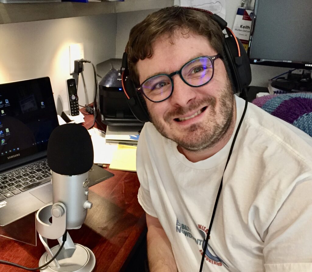  photo of a man in his 30s, with curly light auburn hair, green eyes, a closely trimmed beard and light skin. He is wearing podcast headphones and a microphone, and is smiling warmly. 